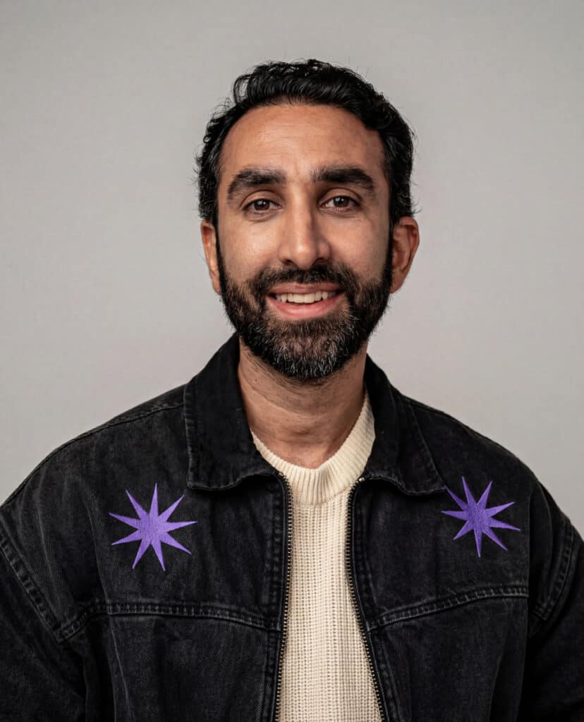 Manick Bhan smiling in a black denim jacket with purple star accents, representing his professional.